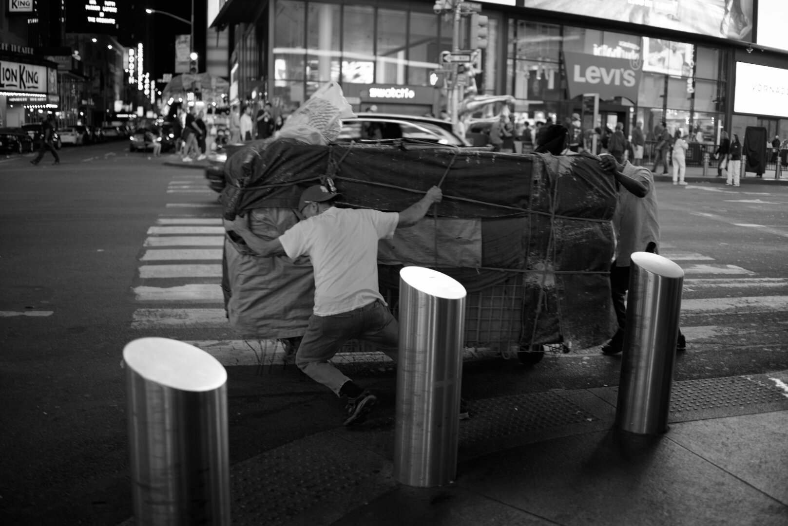 Black and white picture of two guys moving a very large cart of stuff down a street in New York City.