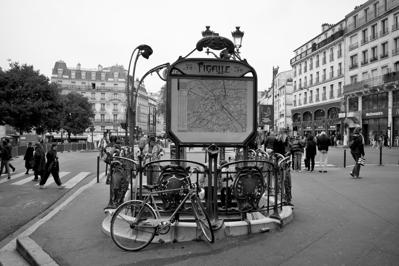 Metro entrance at Pigalle in Paris. Some people milling around. A bike is against a railing. The whole thing would look kind of timeless, except that there is a modern looking McDonald's in the background which really turns the whole picture to crap.