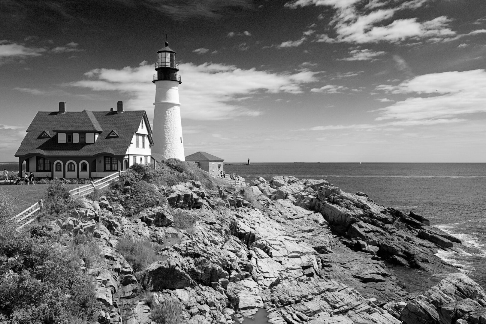 Monochrome pic of a lighthouse with rocks, ocean, and sky around it. And a house. Thje Ram Island Ledge light station is visible across the water.