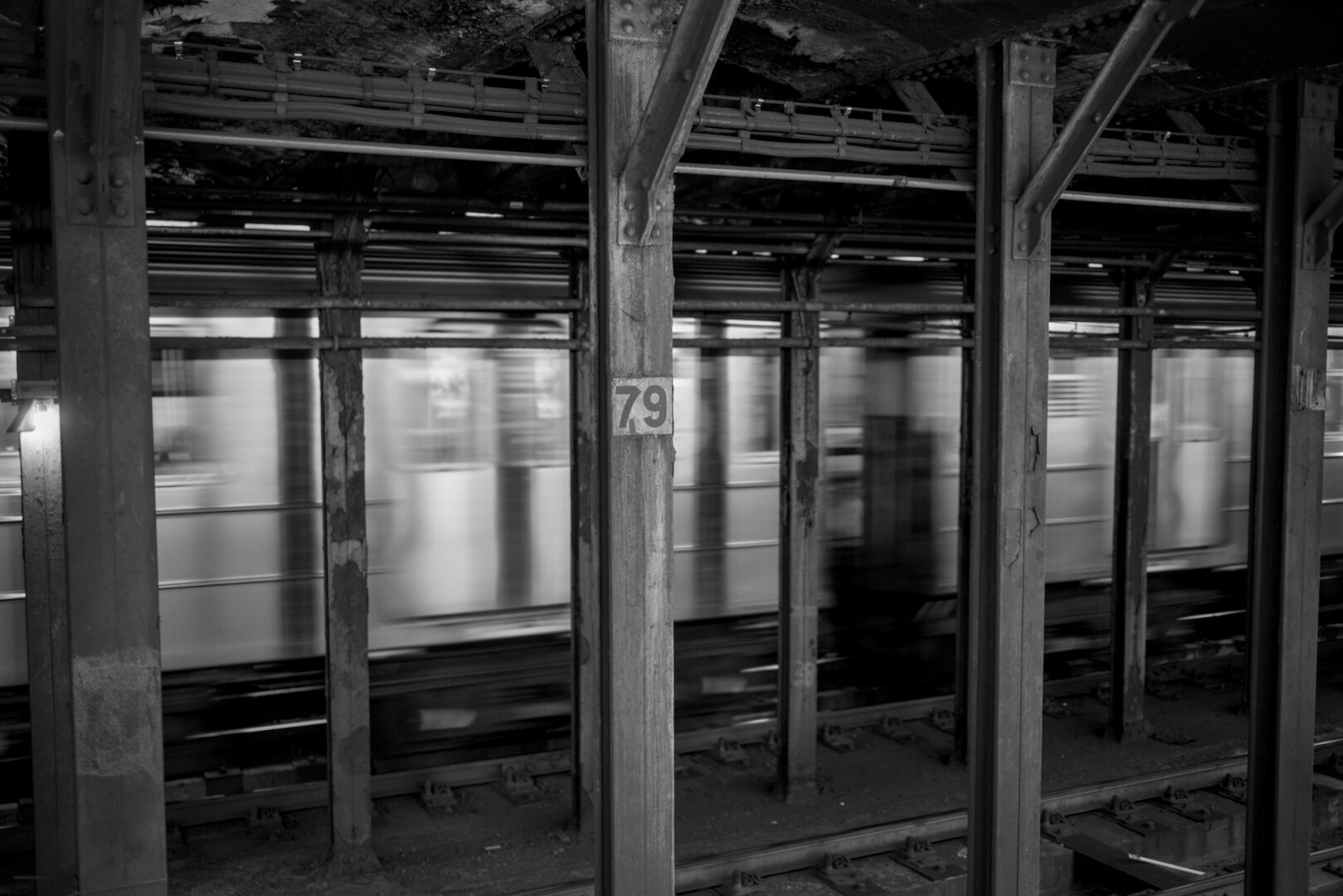 A subway train speeding by several steel support posts. One of the posts has a number 79 on it. The New York subway is dirty and kind of disgusting.