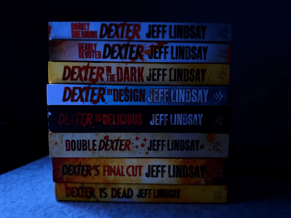 A stack of seven books from Jeff Lindsay's Dexter series, neatly piled on a blue surface against a dark background. From top to bottom, the titles are: "Darkly Dreaming Dexter," "Dearly Devoted Dexter," "Dexter in the Dark," "Dexter by Design," "Dexter is Delicious," "Double Dexter," and "Dexter's Final Cut," with "Dexter is Dead" at the bottom. The spines feature bold red and white text with some blood-splatter designs, reflecting the series' thrilling, crime-filled theme.