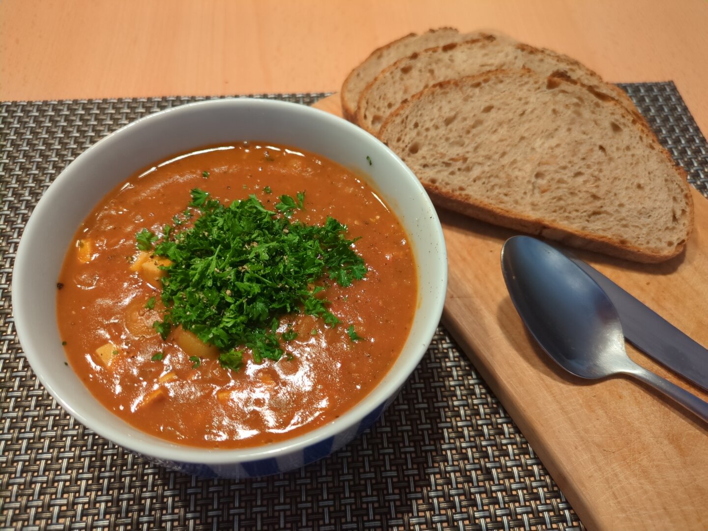 Photograph of a white bowl containing lentil soup topped with parsley. The bowl is located on a dark place mat. Next to it, there's a wooden board with 3 slices of bread, q spoon and a knife. The items are placed on a wooden table.