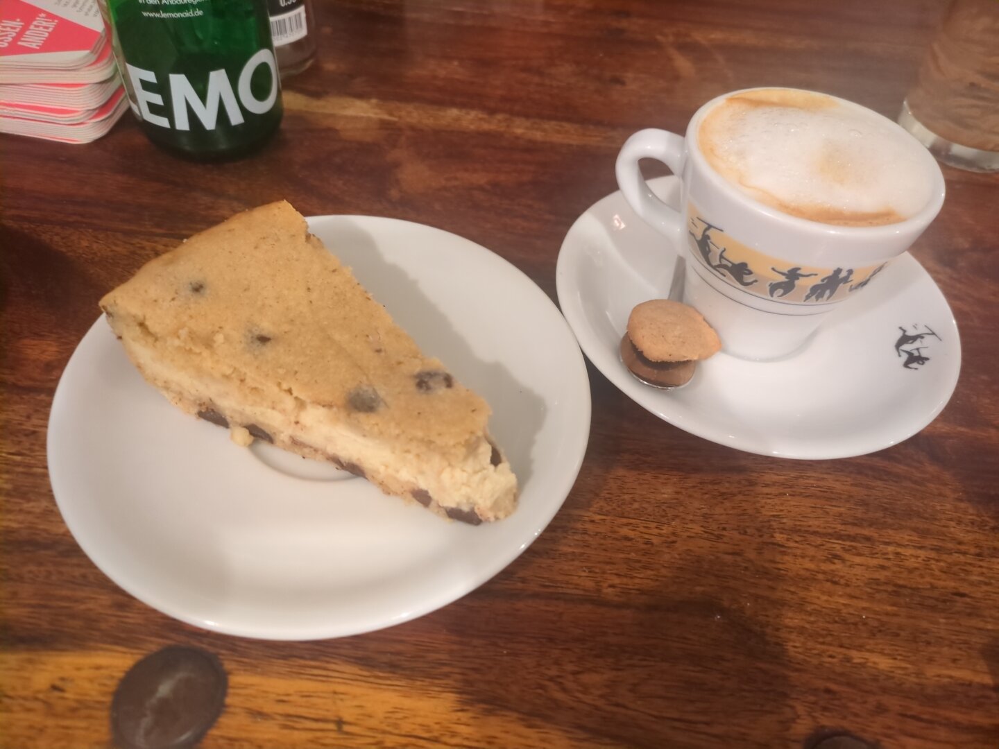 Photograph of a piece of cake on a small white plate, next to it a cup of cappuccino. All items are placed on a wooden table.