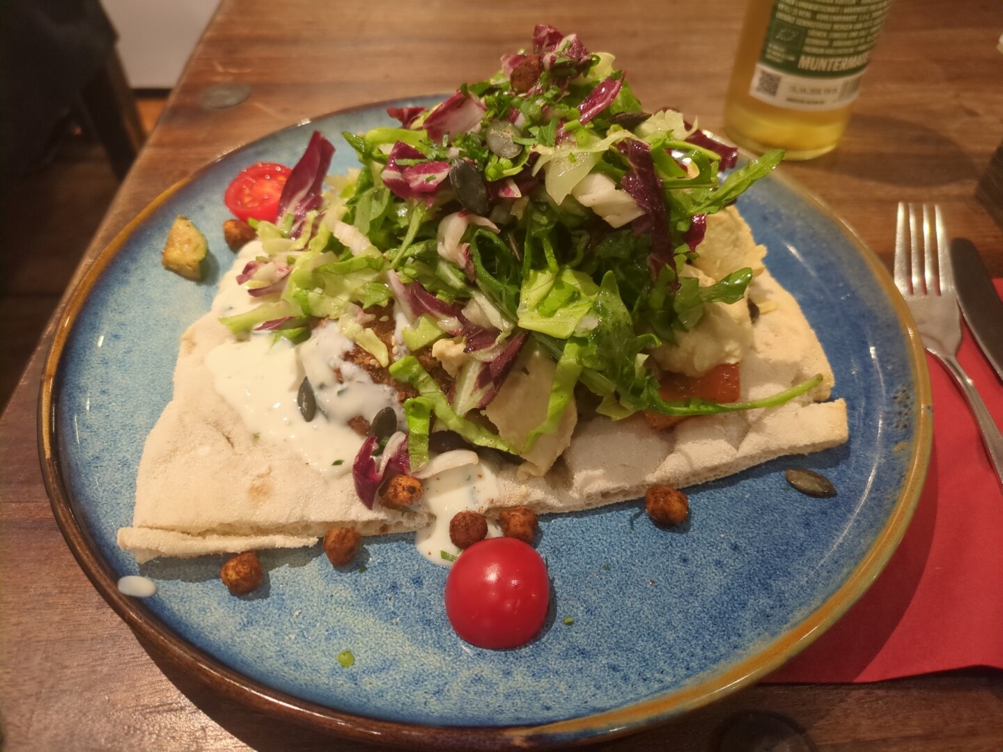 Photograph of a blue plate containing flat bread with a stack of topping, which consists of falafel, humus, (vegan) tzatziki, gilled veggies, and salad. The plate I placed on a wooden table and next to it is a red napkin, cutlery, and a bottle of mate.