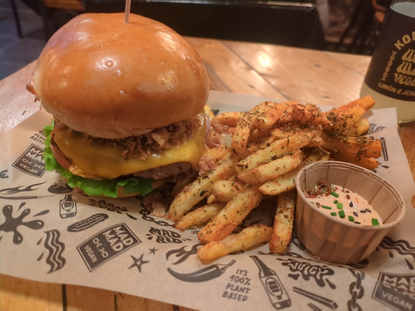 Photograph of a rectangular plate covered with printed paper. On it is a burger, chips and a dip bowl. The plate is on a wooden table and in the background is a kombucha bottle.