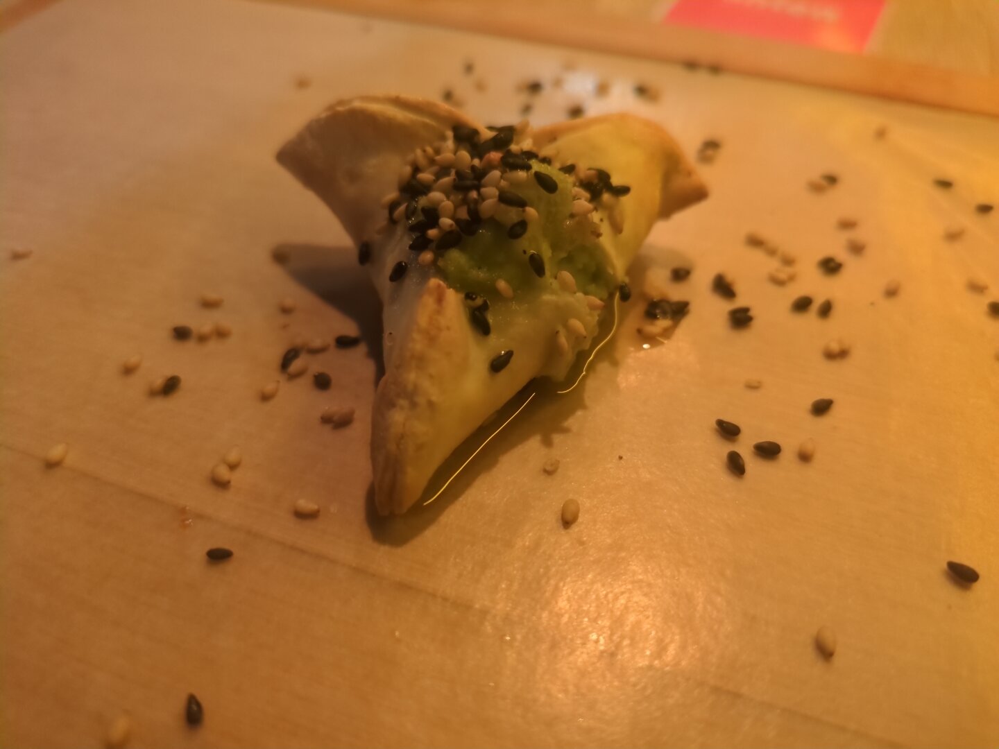 Photograph of a wooden board covered with paper, containing one gyoza with black and white sesame seeds and sprinklers of a spicy oil.