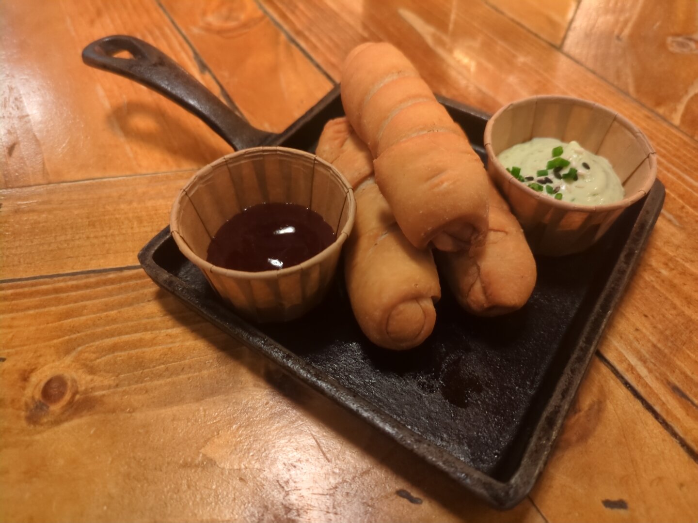 Photograph of 3 (vegan cheese) filled pastry rolls between 2 dip bowls, one filled with red jam, the other with a kind of remoulade. Everything is arranged on a dark square plate, which is placed on a wooden table.