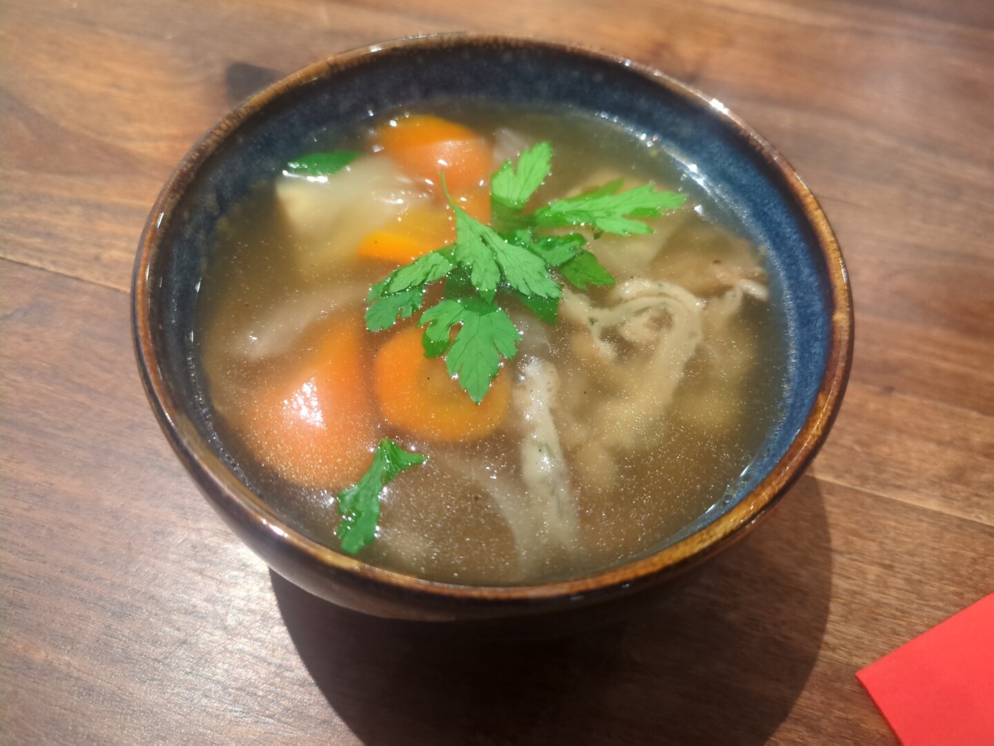 Photograph of a small blue bowl containing the Flädlesuppe, apart from pancake stripes, carrots, onions and parsley are visible in a broth. The bowl is located on a wooden table.