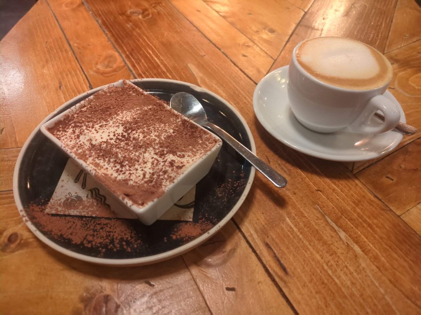 Photograph of a dark round plate with a rectangular white bowl of tiramisu on it, next to it a white cup filled with (oat) milk coffee on a saucer. Everything is on a wooden table.