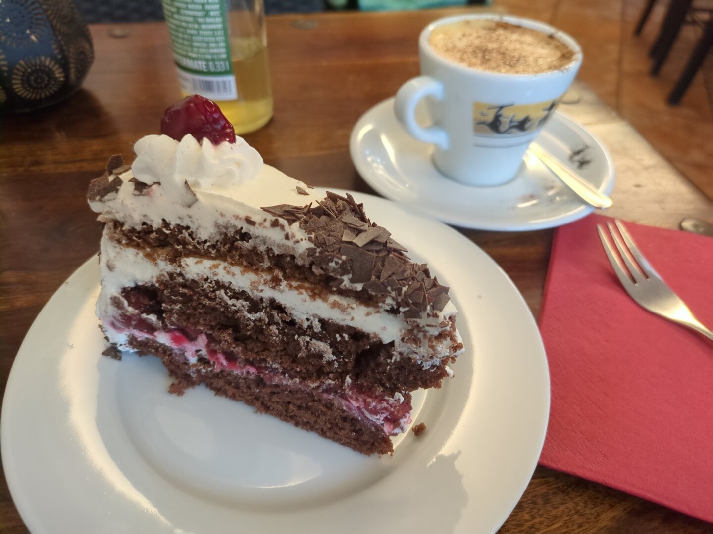 Photograph of a piece of the cake on a small white plate, next to it a cup with cappuccino on a saucer. All items are located on a wooden table. In the background there's a red paper napkin, a fork and a bottle of mate visible.