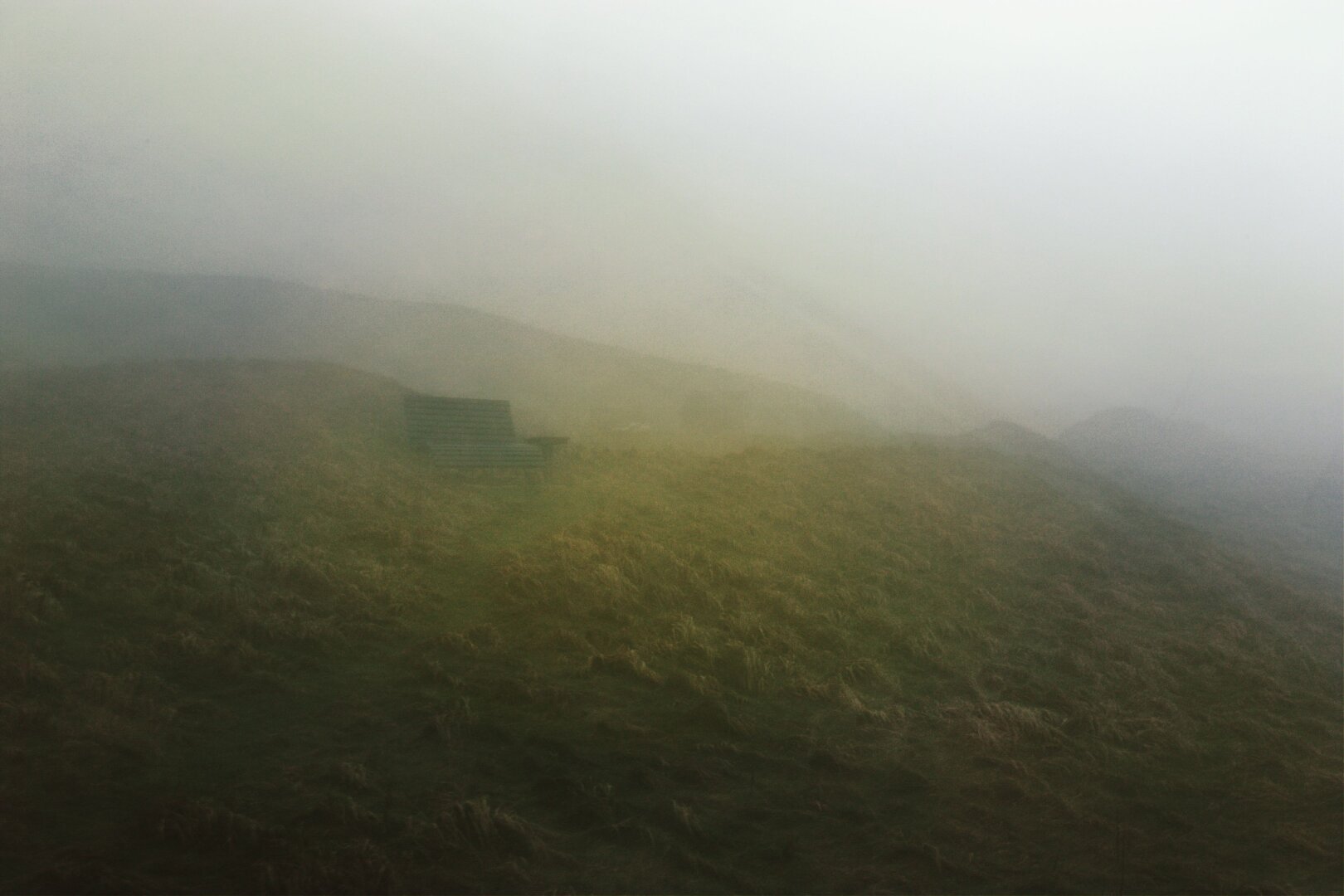 Wooden bench in the fog. Green hills in front, beside and behind. All around it.