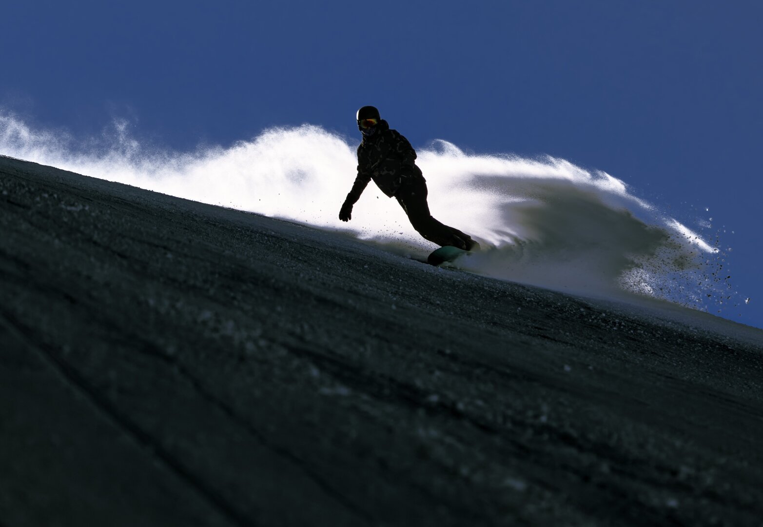 Snowboarder plunges down the slope with a snow fountain illuminated from behind.