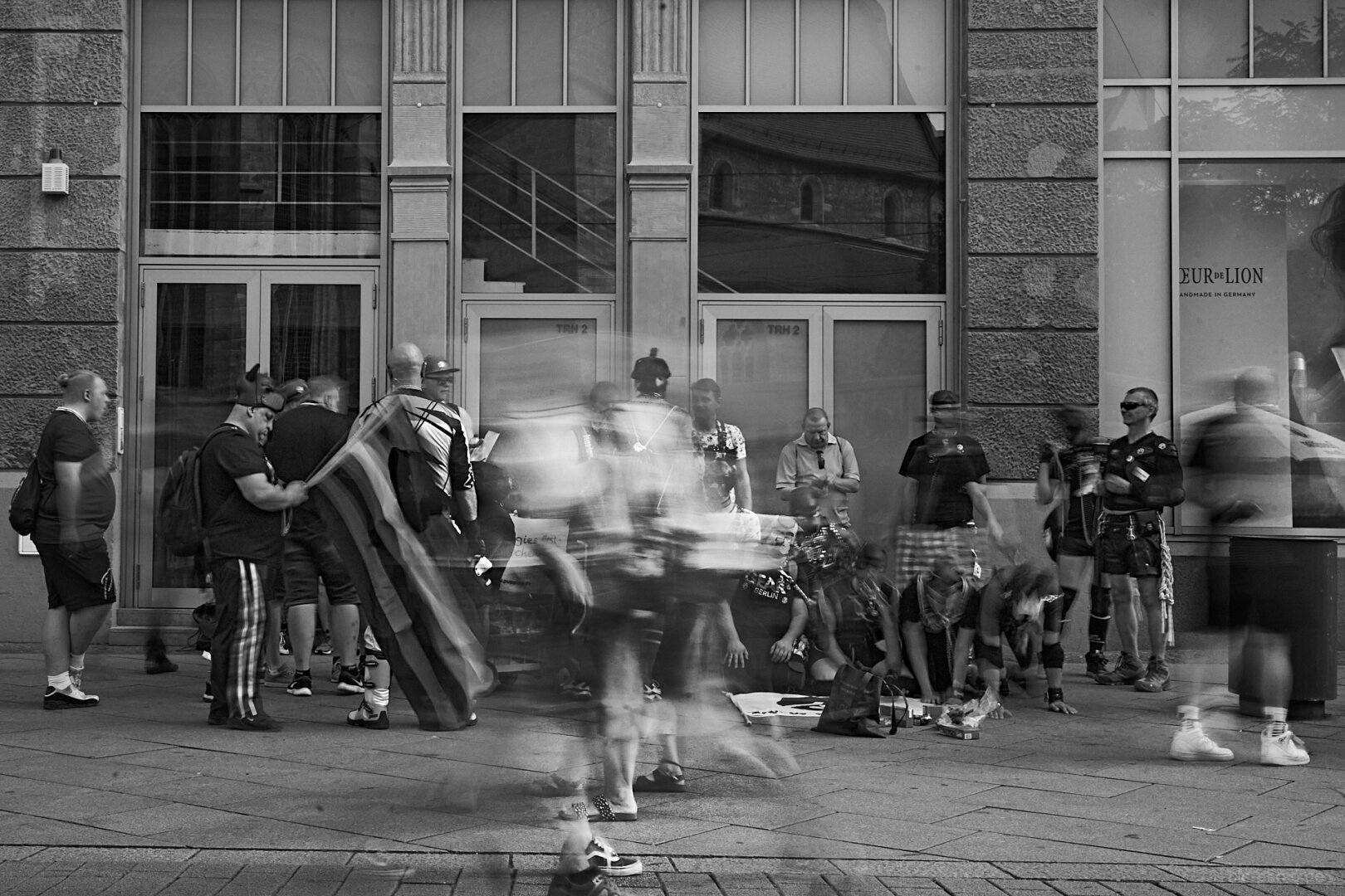 A black-and-white street photograph captures a busy scene outside a large stone building. A group of people gathers along the sidewalk, while several passersby are blurred by motion, creating a strong sense of movement. Some people appear to be wearing distinctive costumes or traditional-looking outfits, and one person on the right wears sunglasses and a leather-style outfit.

The building behind them has tall multi-pane windows, ornate stone detailing, and large glass doors. Reflections in the windows add another layer of visual detail. The contrast between the sharply defined architecture and the ghostlike motion blur of the pedestrians gives the photograph a dynamic, almost cinematic quality.