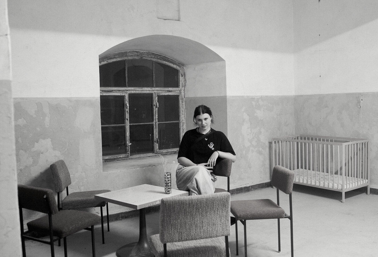 This black-and-white photo shows a young woman sitting alone on a chair in a sparsely furnished room with peeling walls. The room contains a small table surrounded by four chairs and a baby crib in the corner. A large, old window with multiple panes is behind her, adding to the vintage and somewhat austere atmosphere. (Aria)