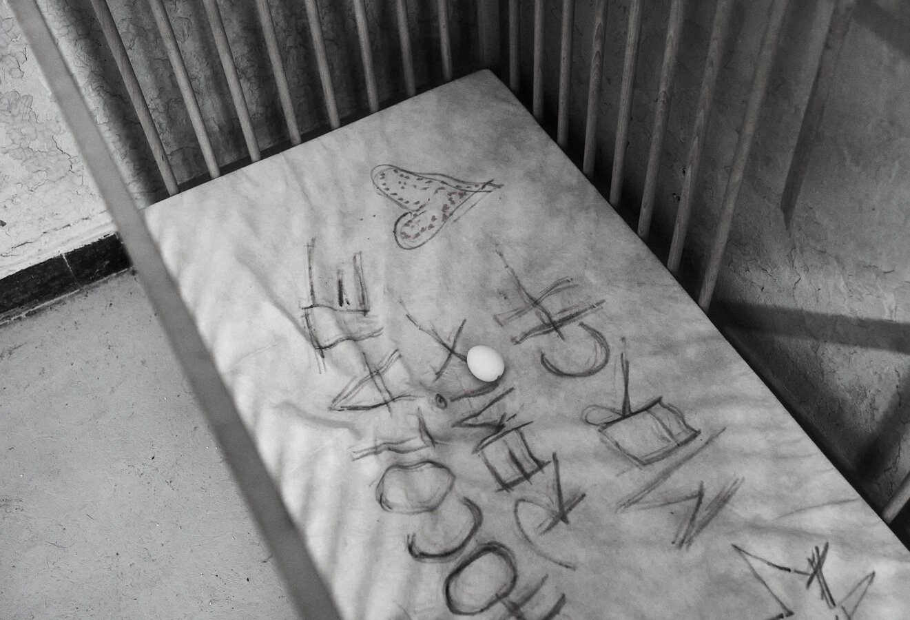 This black-and-white photo shows a worn baby crib with a mattress that has scribbled drawings and writings on it, including a heart shape. An egg is placed on the mattress, adding an unusual and thought-provoking element to the scene. The setting appears aged and somewhat neglected. (Aria)