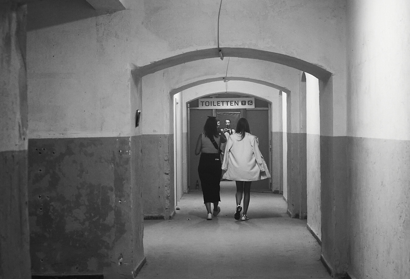 This black-and-white photograph shows two women walking down a long, narrow corridor with arched ceilings and worn walls. At the end of the corridor, there is a sign above a door that reads "TOILETTEN," indicating restrooms. The women are seen from behind, and their reflections, along with a man taking the photo, are visible in a mirror or glass panel near the door. The image captures a candid, somewhat vintage or timeless atmosphere. (Aria)