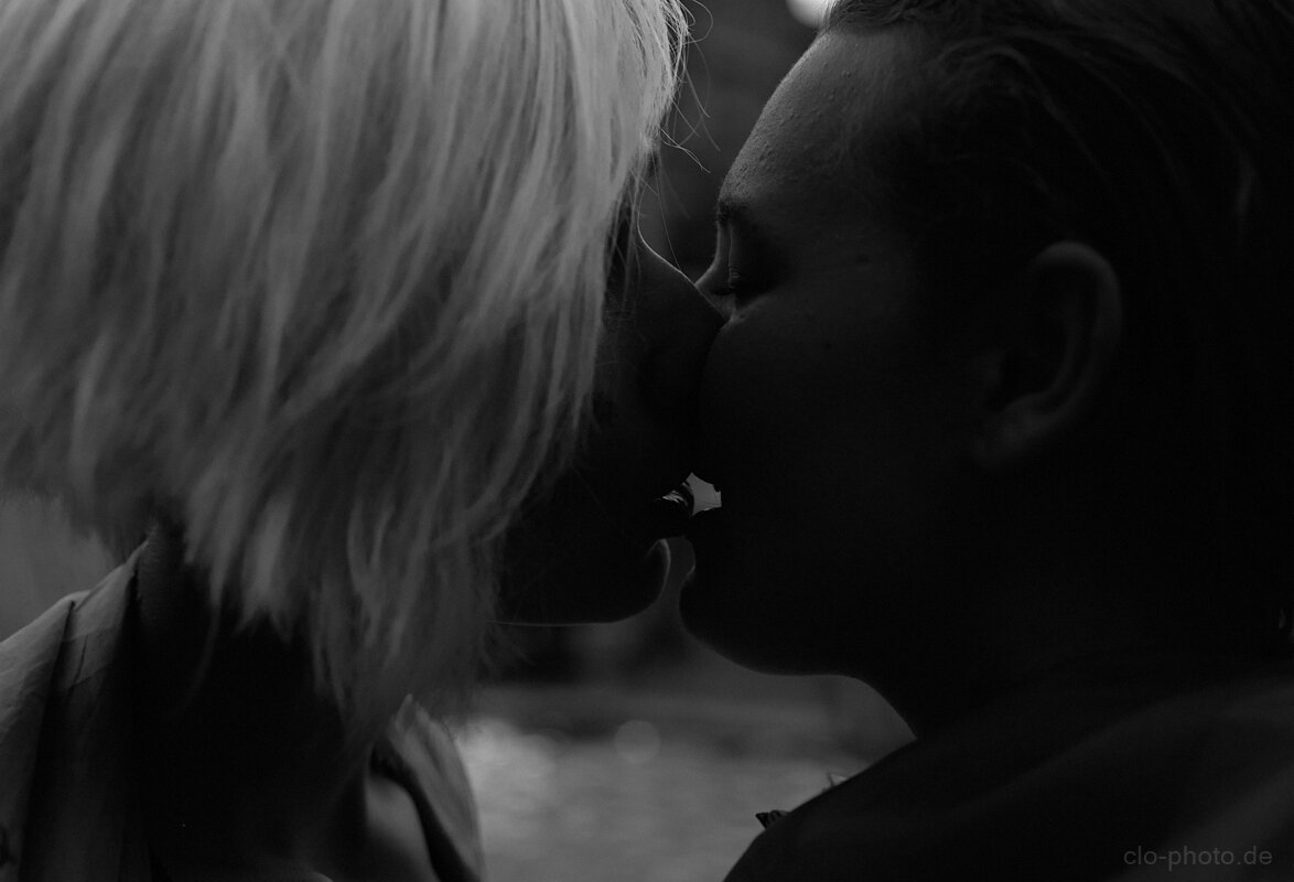 A black-and-white close-up photograph captures two people kissing in profile. Their faces are largely silhouetted against a softly blurred background, with the lighting emphasizing the contours of their noses, lips, cheeks, and foreheads.

One person has light, shoulder-length hair that fills much of the left side of the frame, while the other has short, dark hair and occupies the right side. Their embrace is intimate and quiet, with a hand or arm appearing around the other person’s neck.

The shallow depth of field and deep shadows create a tender, cinematic mood, while the tight framing makes the kiss the unmistakable focal point.