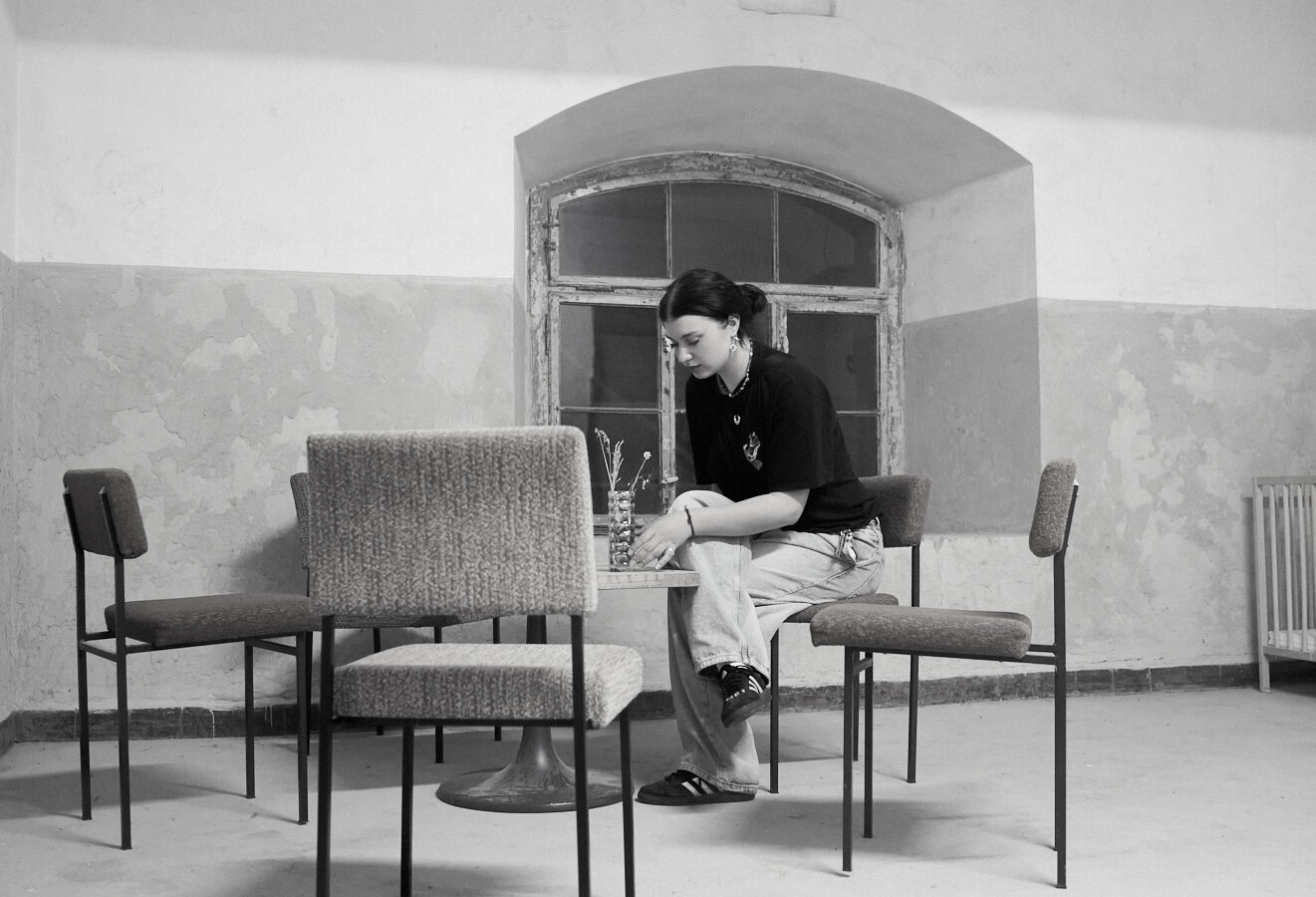 This black-and-white photo depicts a young woman sitting alone at a small table in a sparsely furnished room with peeling walls. She is focused on a vase with dried flowers on the table. The room features an old, arched window and a baby crib in the corner, creating a quiet, contemplative atmosphere. (Aria)