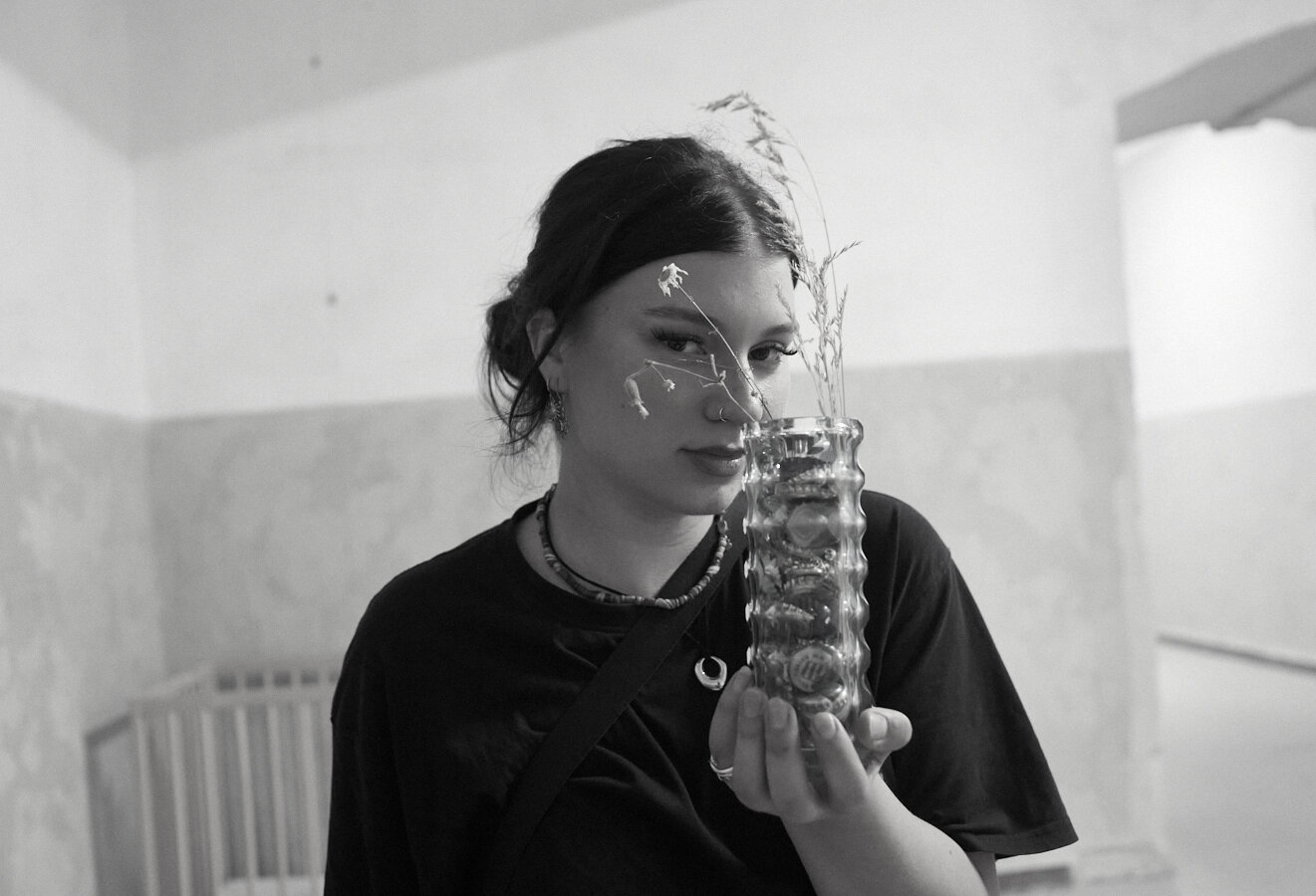 This black-and-white portrait shows a young woman holding a textured glass vase with dried flowers close to her face. She gazes softly at the camera, with a worn wall and a baby crib visible in the background, creating an intimate and contemplative mood. (Aria)