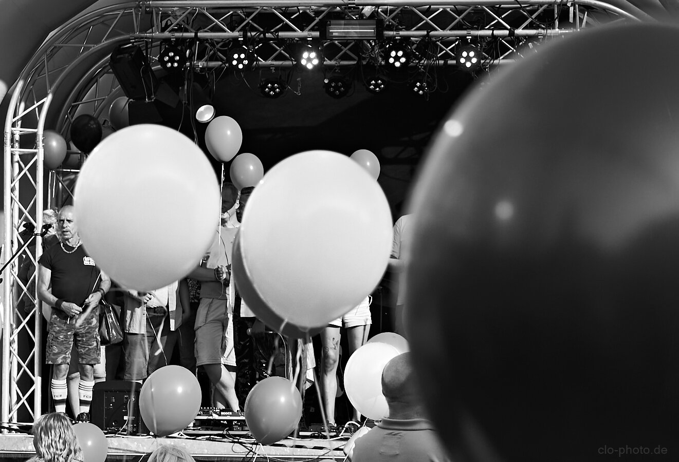 A black-and-white photograph shows a lively outdoor stage scene filled with large balloons. Several performers or participants stand on a small stage beneath a curved canopy and a framework of metal trusses supporting numerous stage lights.

Large balloons dominate the foreground, partially obscuring the people behind them and creating a layered, playful composition. On the left, an older woman stands near the edge of the stage, while several other people are visible between the balloons. In the lower foreground, the backs of spectators’ heads frame the scene.

The strong monochrome contrast emphasizes the glowing balloons, dark stage area, and bright overhead lights. A large out-of-focus balloon or object occupies much of the right side, giving the image an intimate, immersive viewpoint from within the crowd.