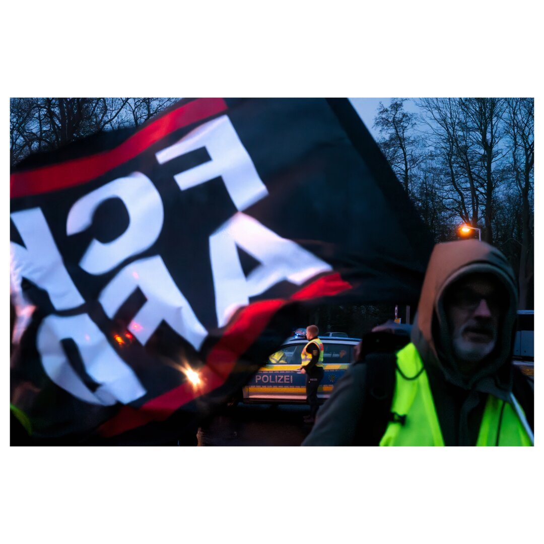 A documentary photograph of a protest scene at dusk, showing flags, high-visibility vests, and police presence in the background