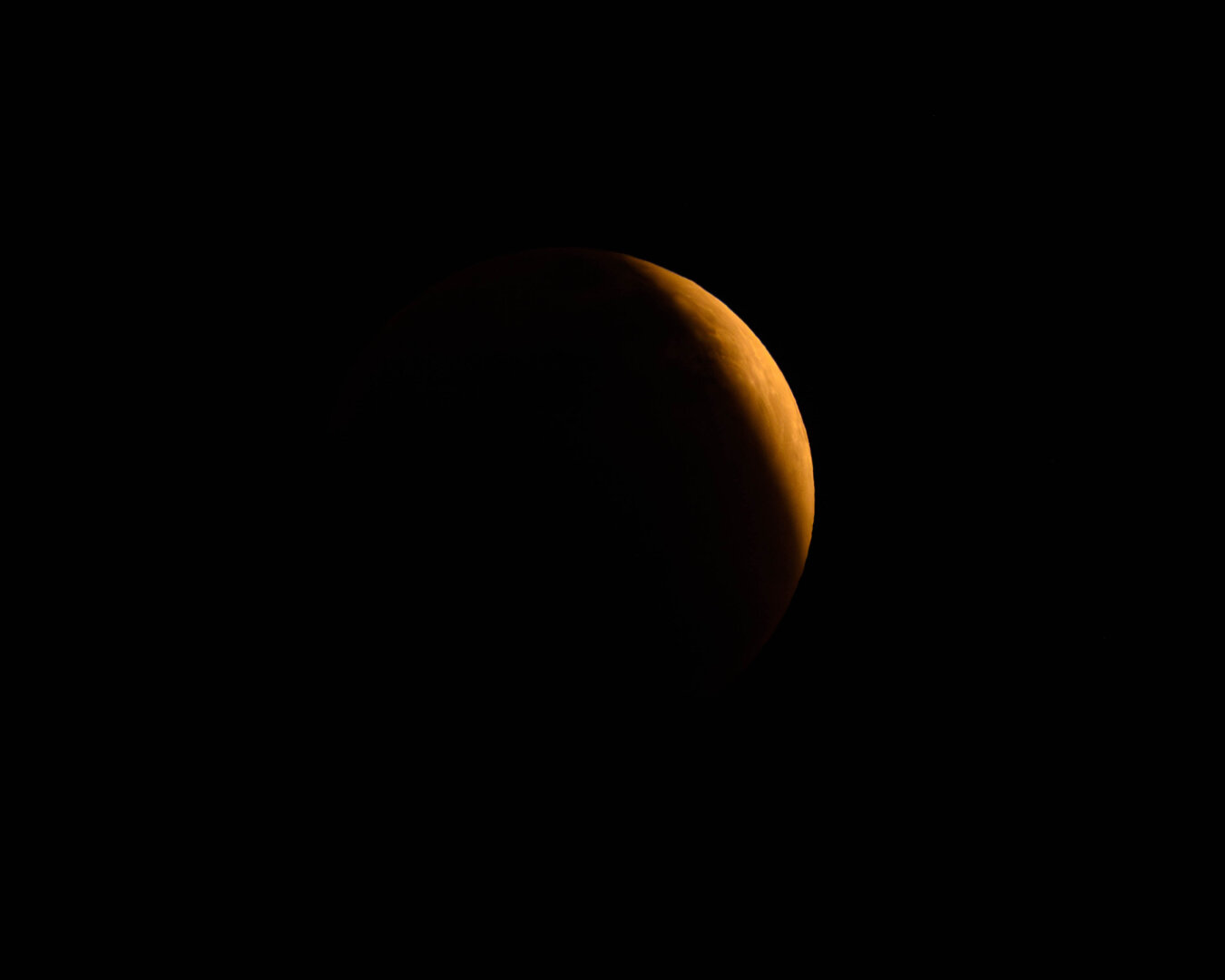 A partially illuminated view of the moon against a black background, featuring a reddish-orange hue during the lunar eclipse.