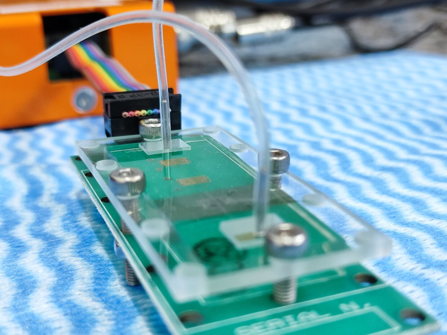 Foto de uma placa de circuito eletrônico impresso, possuindo pequenos eletrodos interdigitados. Ao fundo, se observa microcanais fluídicos (tubinhos) transparentes, que passam por cima da placa.