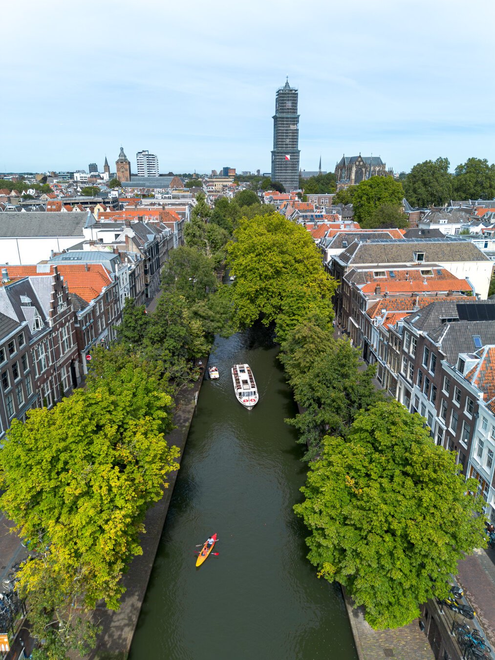 Drone eye view of the Oude Gracht in Utrecht. With green trees lining the canal and some church towers in the background.

In the canal you can see a red canoe and a white tourist boat.