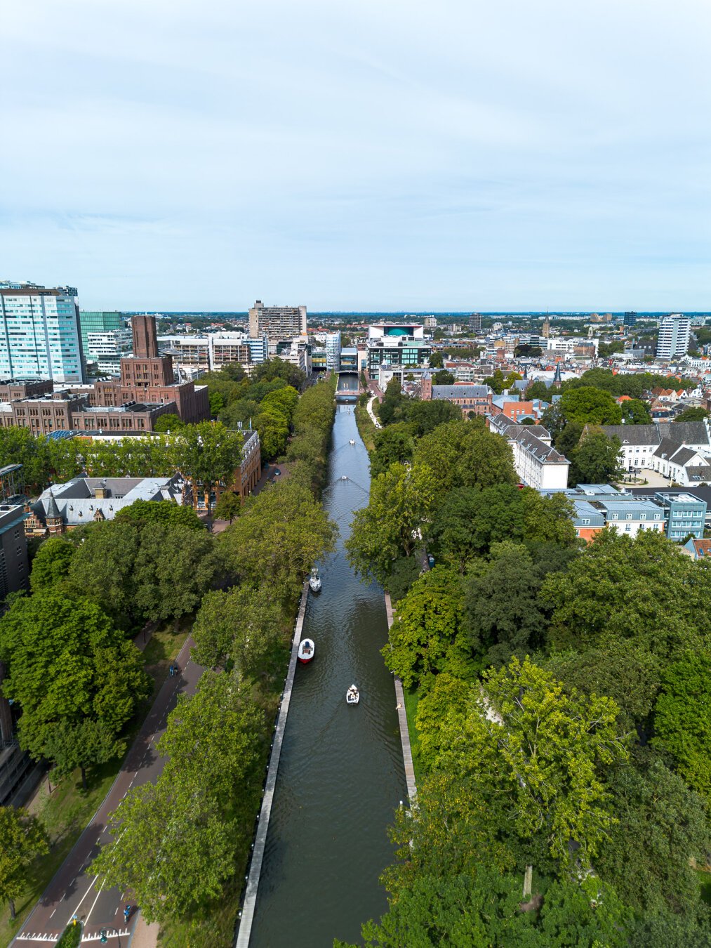 Drone shot of a restored canal in Utrecht, the Netherlands. Long stretch of water in the center, lined by trees, and in the near distance a city scape with some medium and high rises.