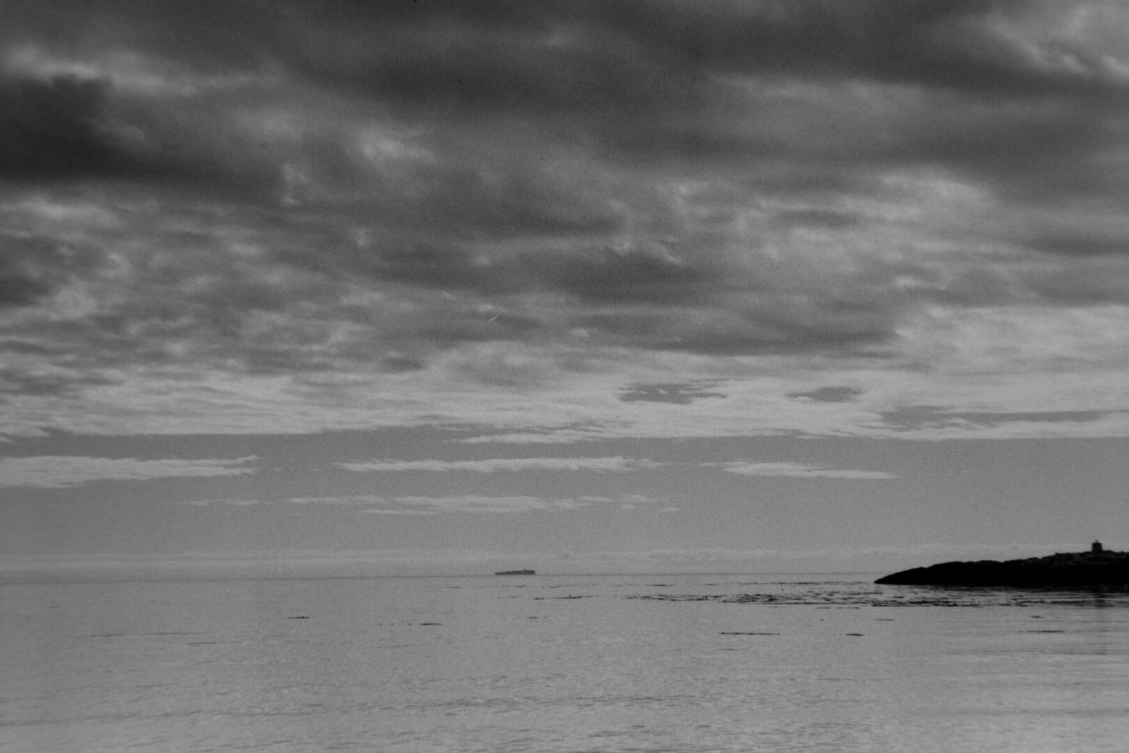 A view from the beach shore. Out over the water a dark sky looms above a low island.
