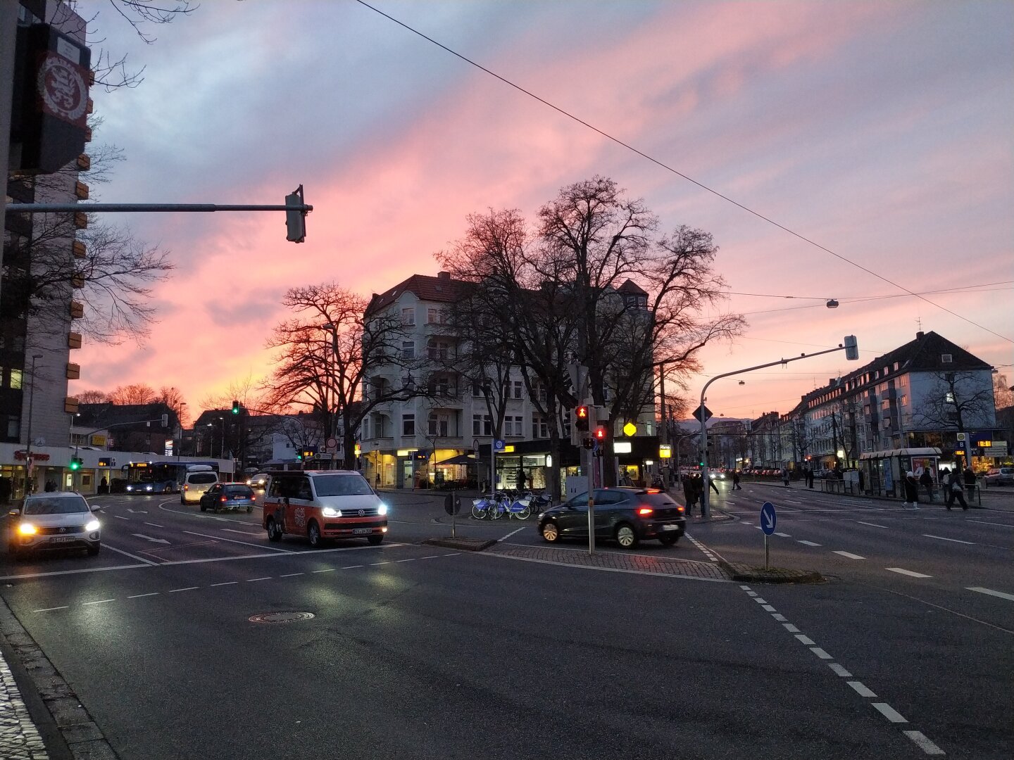 Straßenkreuzung im Zwielicht. Blauer Himmel mit rosa Wolken. Die Fahrzeuge haben schon die Lichter an. Die Bäume im Hintergrund sind kahl.