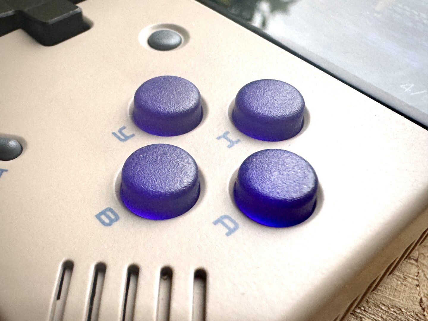 Close-up view of a gaming handheld with four circular buttons arranged in a diamond pattern. The buttons are labelled with letters: B, A, Y, and X. The buttons have a purple hue and a subtle texture.