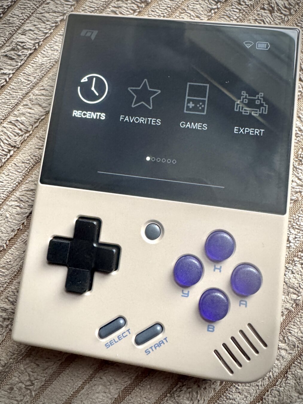 A handheld gaming device with a retro design reminiscent of a Game Boy. It features a black and cream colour scheme with a large screen on the upper half displaying icons labelled "Recents," "Favorites," "Games," and "Expert." The lower half has a control pad on the left, four circular purple buttons on the right marked "Y," "X," "B," and "A," and two smaller buttons below marked "Select" and "Start."