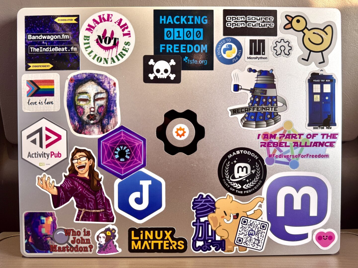 The lid of a silvery 14 inch Framework laptop covered in stickers, including the logos of many #Fediverse-related technologies and platforms. There is a cluster of Mastodon stickers in the bottom right corner of the case.