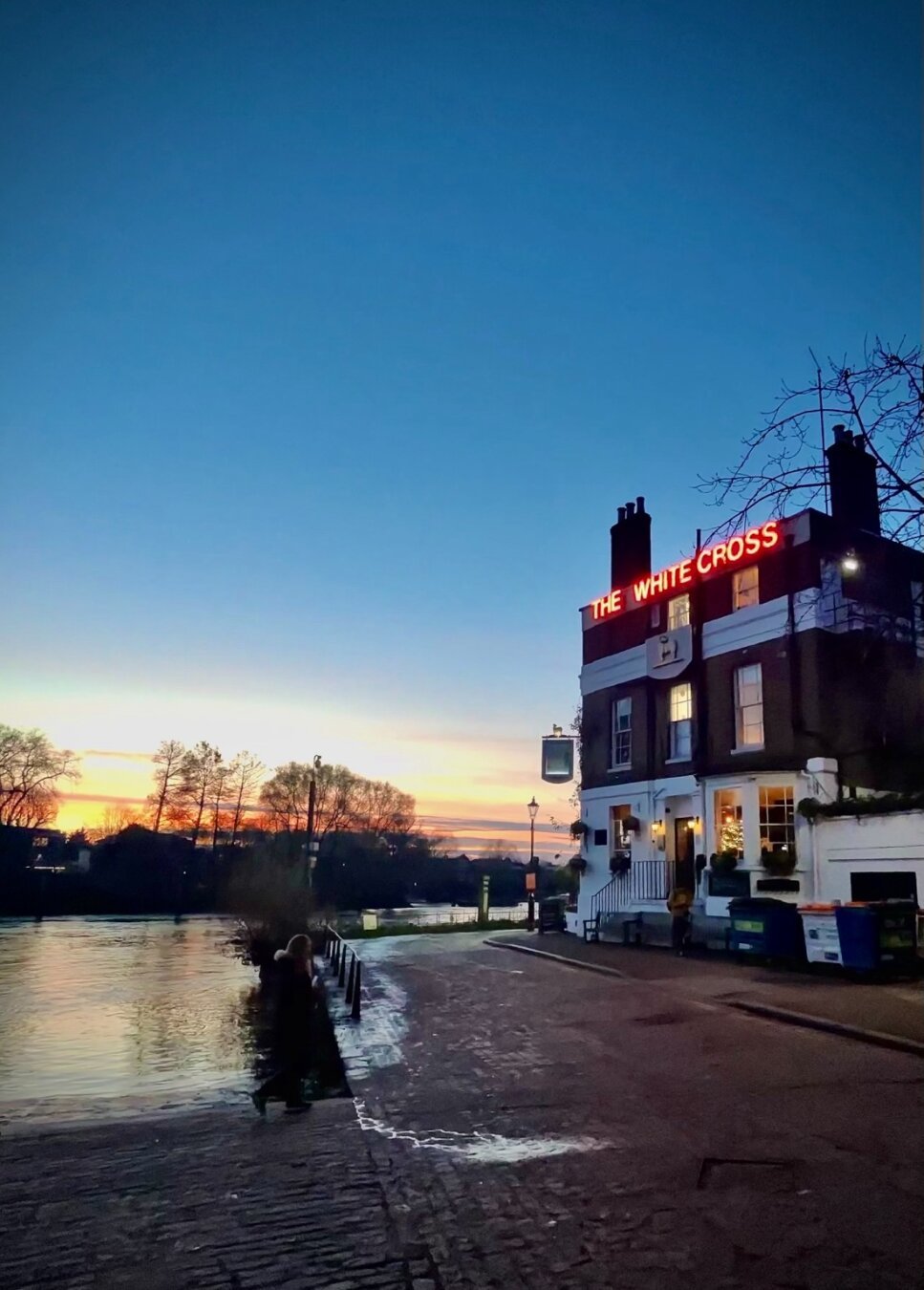 Looking towards the sunset at the edge of the river. The water laps at a ramp to a cobbled street in the front left corner. A pub stands to the right, a tall square building with a red neon sign announcing the name The White Cross. The sun has nearly set beyond dark trees over the water.