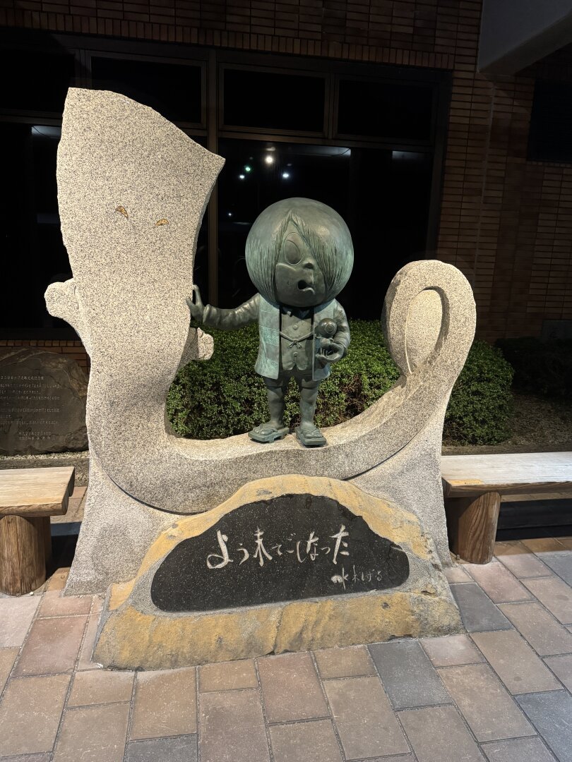 A statue of a small confused looking man standing on a bench. In his left hand stands another small character. There is Japanese text on a plaque in front: ようれでしたった