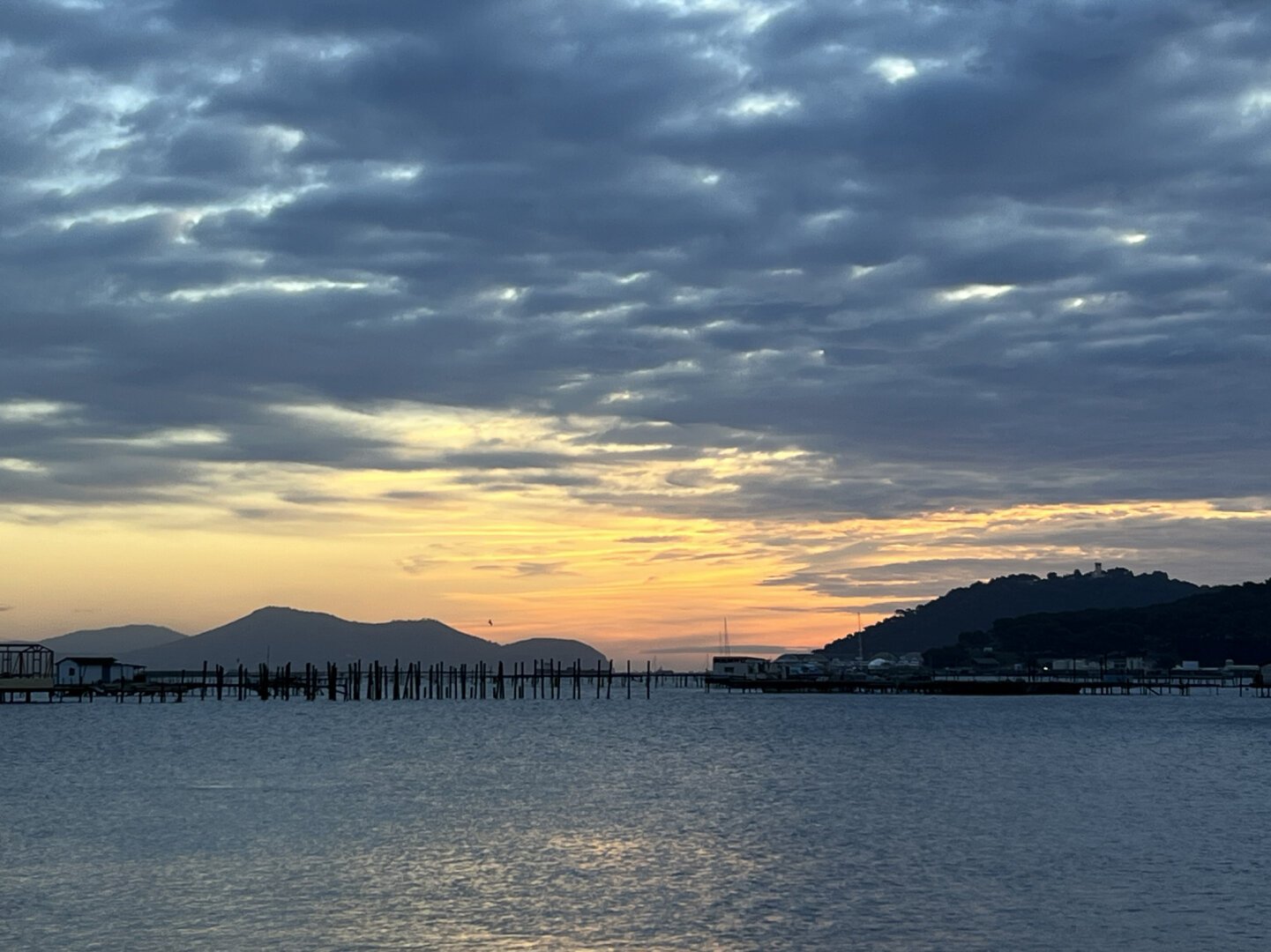 Blue seascape and blue grey clouds, with black fishing structures in the harbor and yellow orange twilight behind black little mountains