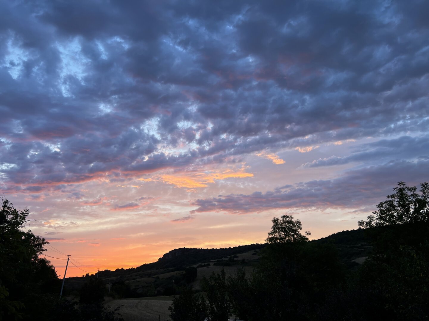 Encore une photo de paysage crépusculaire, un peu plus tard que la précédente et d'un autre point de vue. Beau dégradé dans le ciel, de l'orange à l'horizon dégagé au bleu gris dans les nuages. Ligne de crête obscure, bien contrastée, avec quelques arbres au premier plan et des champs derrière.