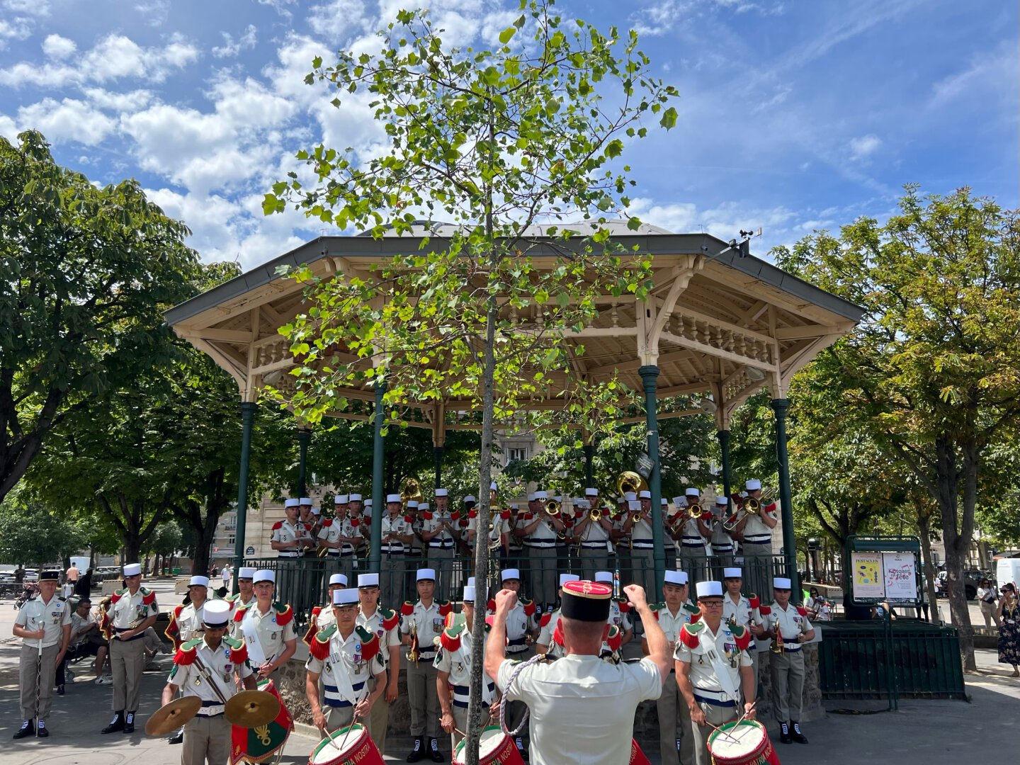 Légionnaires en képi blanc, épaulettes rouges et uniforme beige, jouant de leurs instruments dans le vieux kiosque et juste devant. Tambours, cimballes et instruments à vent.