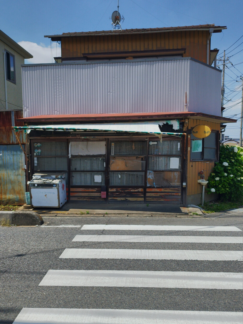 At the end of a crosswalk stands a two-storey structure, decades old. The top is residential and appears to be still occupied. The bottom level was a store with sliding doors, an old cooler still sits to the left of those. All curtains are drawn and the elements have gone to work on the buildings's wood. The awning has been shredded by wind. Taken on a sunny day.