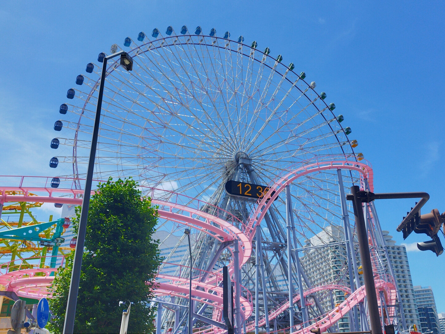 A slightly skewed look up at Yokohama's famous ferris wheel. In front of it are the pink tracks of a rollercoaster and a tree. The sun is shining at 12:36pm according to the massive digital clock on the wheel.