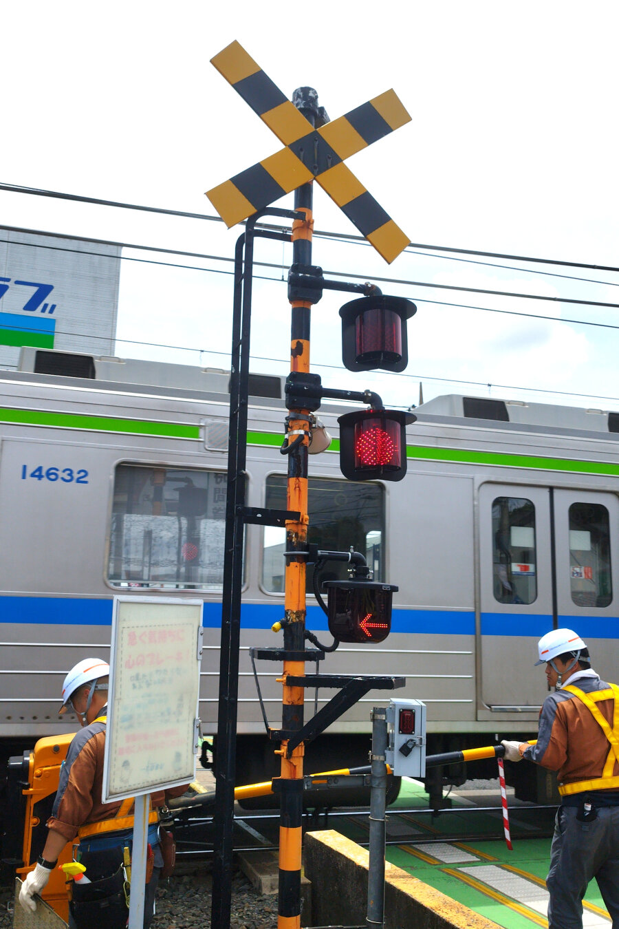 Portrait shot of the railroad crossing sign next to the station. The Japanese version is a saltire of black-and-yellow dashed lines on a yellow pole. Underneath are dual blinking lights so a least one might work even if the other gave up the ghost. And further below that are indicator arrow lights to show the direction the train is conning from. It shows from the right here. The black and yellow barriers are lowered and the gray commuter train is already crossing and we see a sliver of it in the background. There are two men of a maintenance crew in hardhats and high vis uniforms also in the shot. One is looking at the pole that is raising and lowering the barrier and another is inspecting the cross barrier itself. It's sunny.