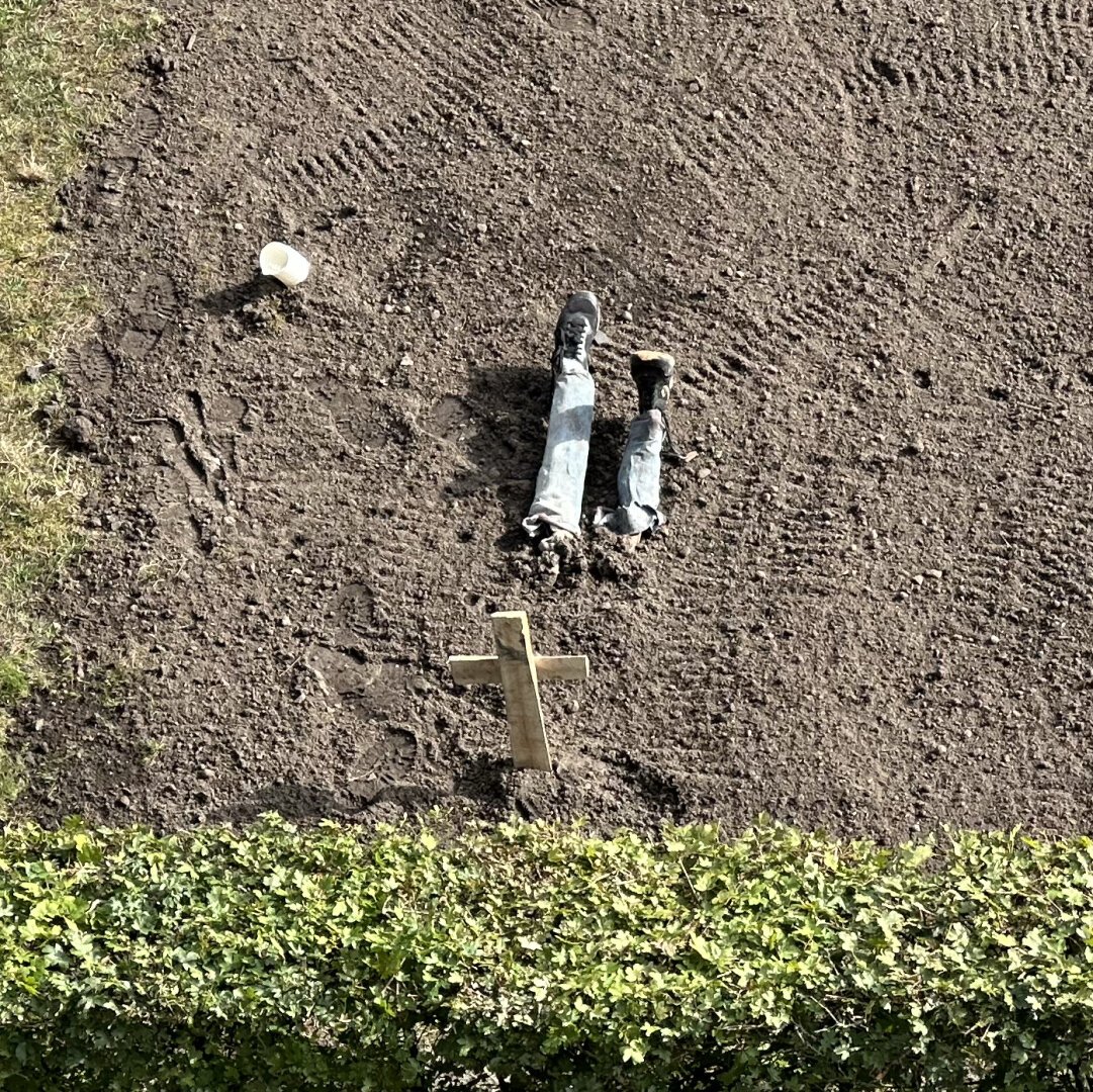 Fake lower legs sticking out of freshly dug ground with a wooden cross on the other side insinuating somebody ended up in a shallow grave.