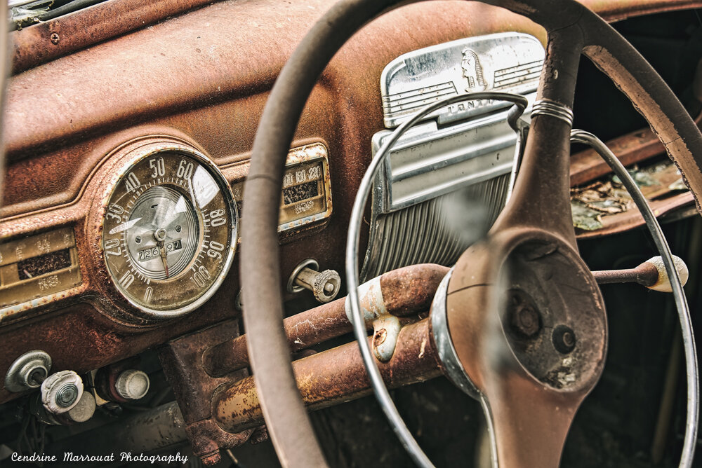 The dashboard of an old car, probably from the 1950s or 1960s. It is rusty and the spinner knob of the steering wheel is missing.