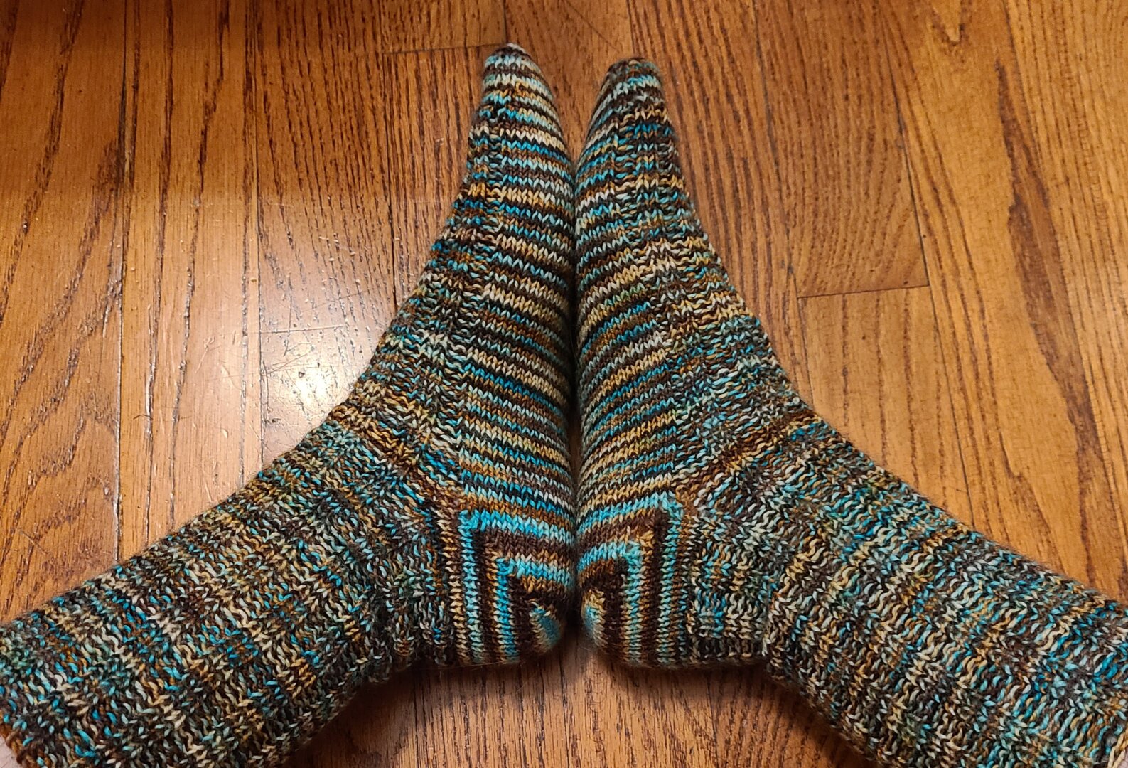 A pair of feet with the soles pressed together wearing hand knit socks in mottled stripes of teal, brown, and gold.