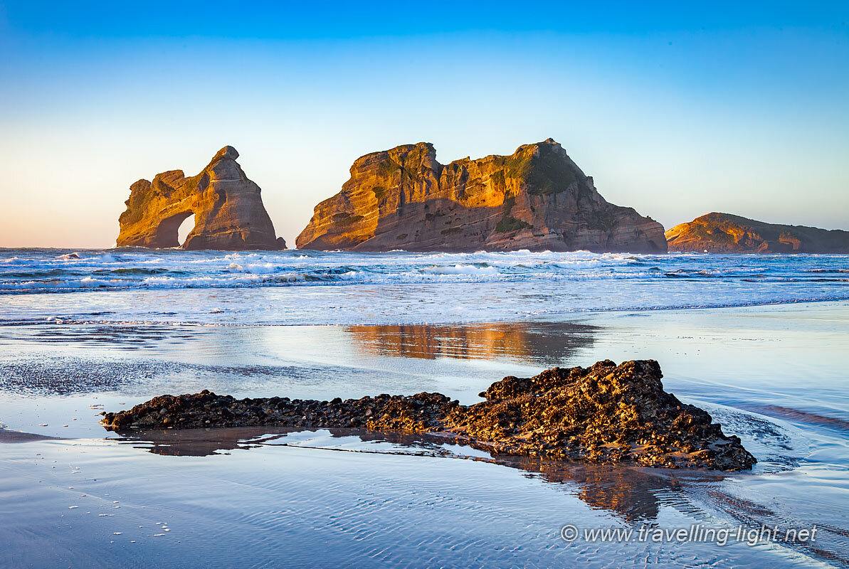 Photo of a flat sandy beach on a clear evening with incoming tide washing around a small rock outcrop in the foreground, and two tall rocky islands, one with an arch, in the middle distance.