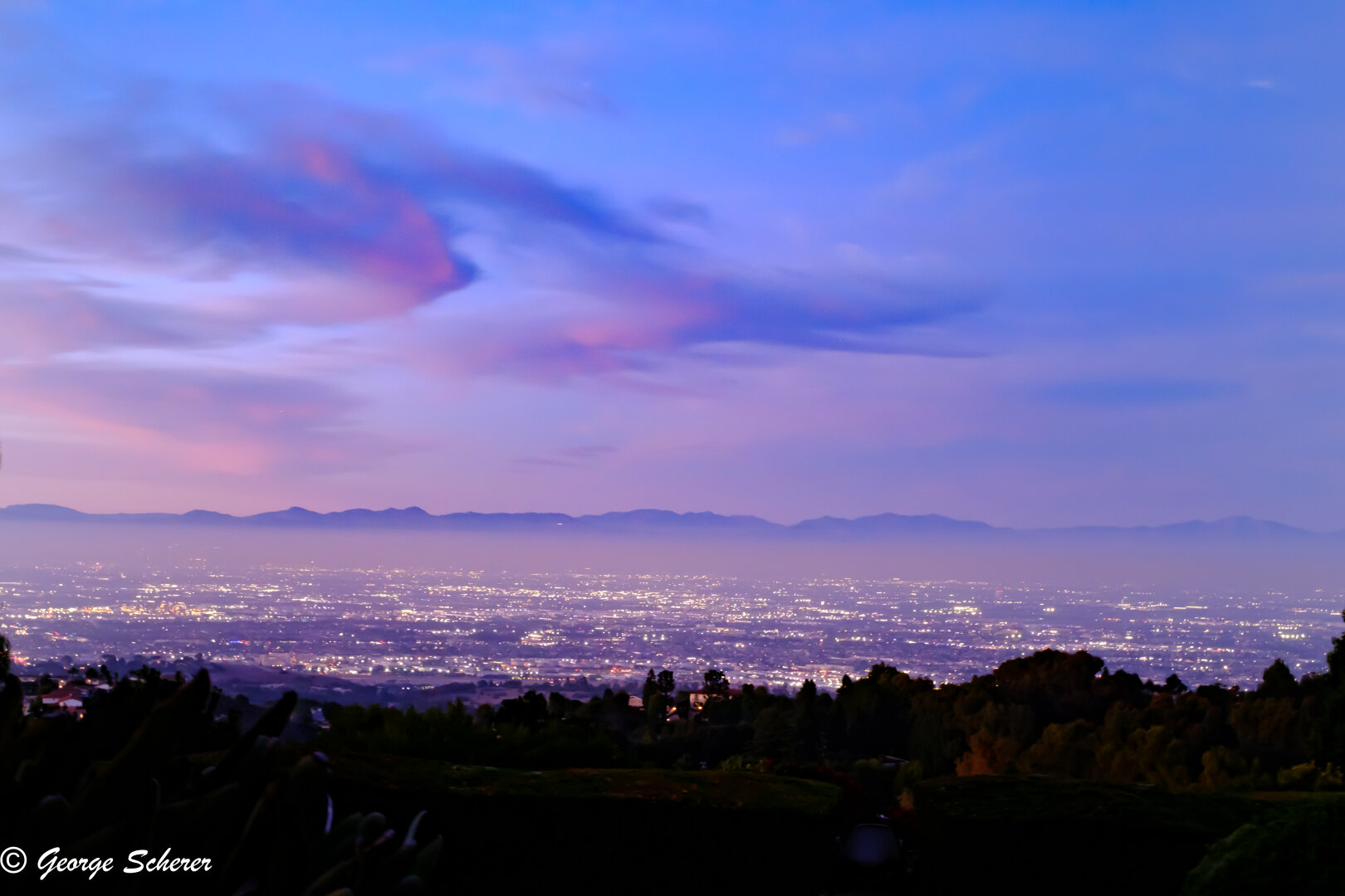 View of the lights in the LA basin, with a layer of fog, and the mountains in the background.  Sunset has lit up the clouds in colors of dark blue and pink.