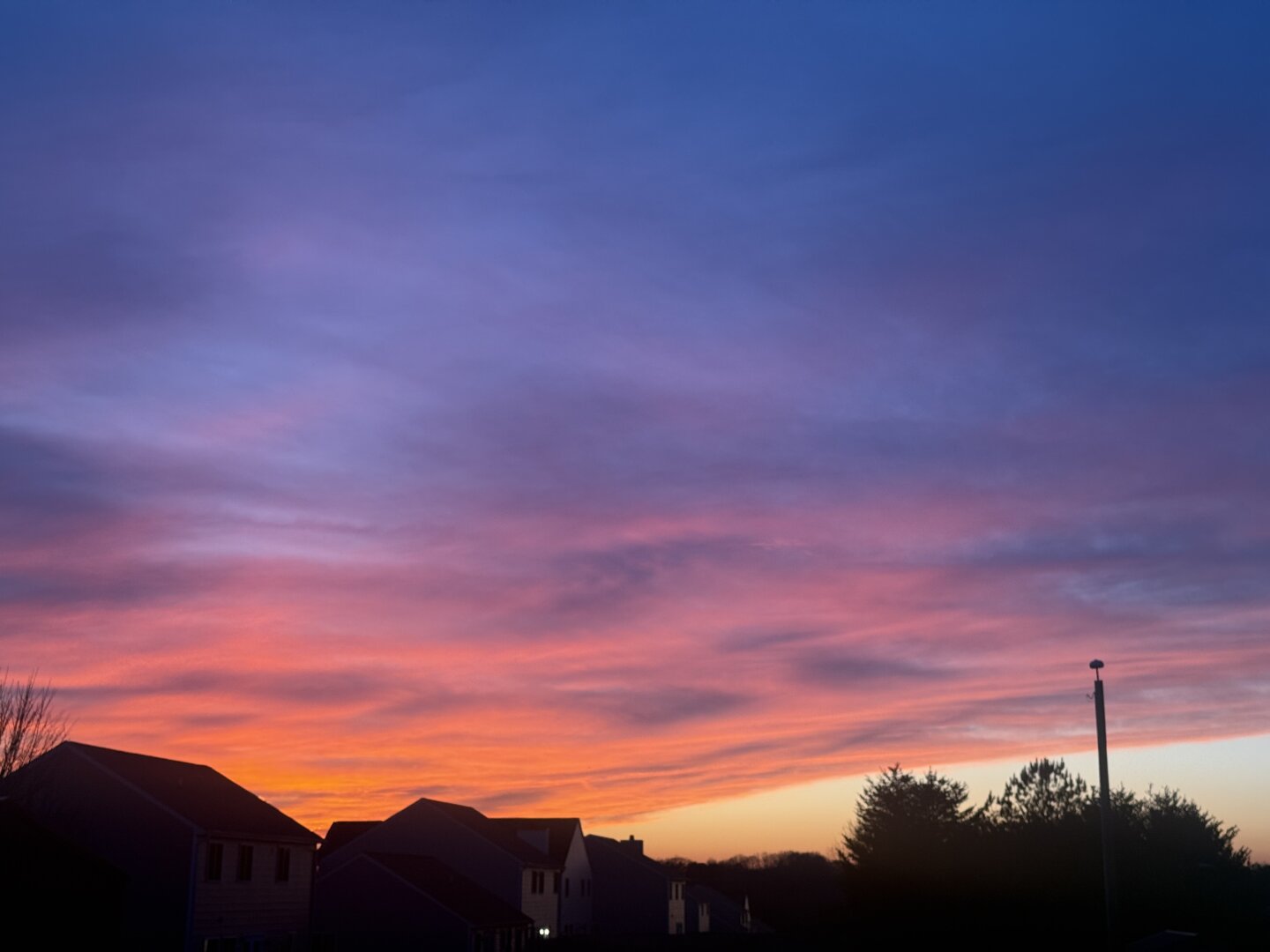 A vibrant sunset sky displays hues of orange, pink, and purple, with thin clouds streaked across it.  Silhouetted against the sunset are several houses, some trees, and a tall, thin pole.