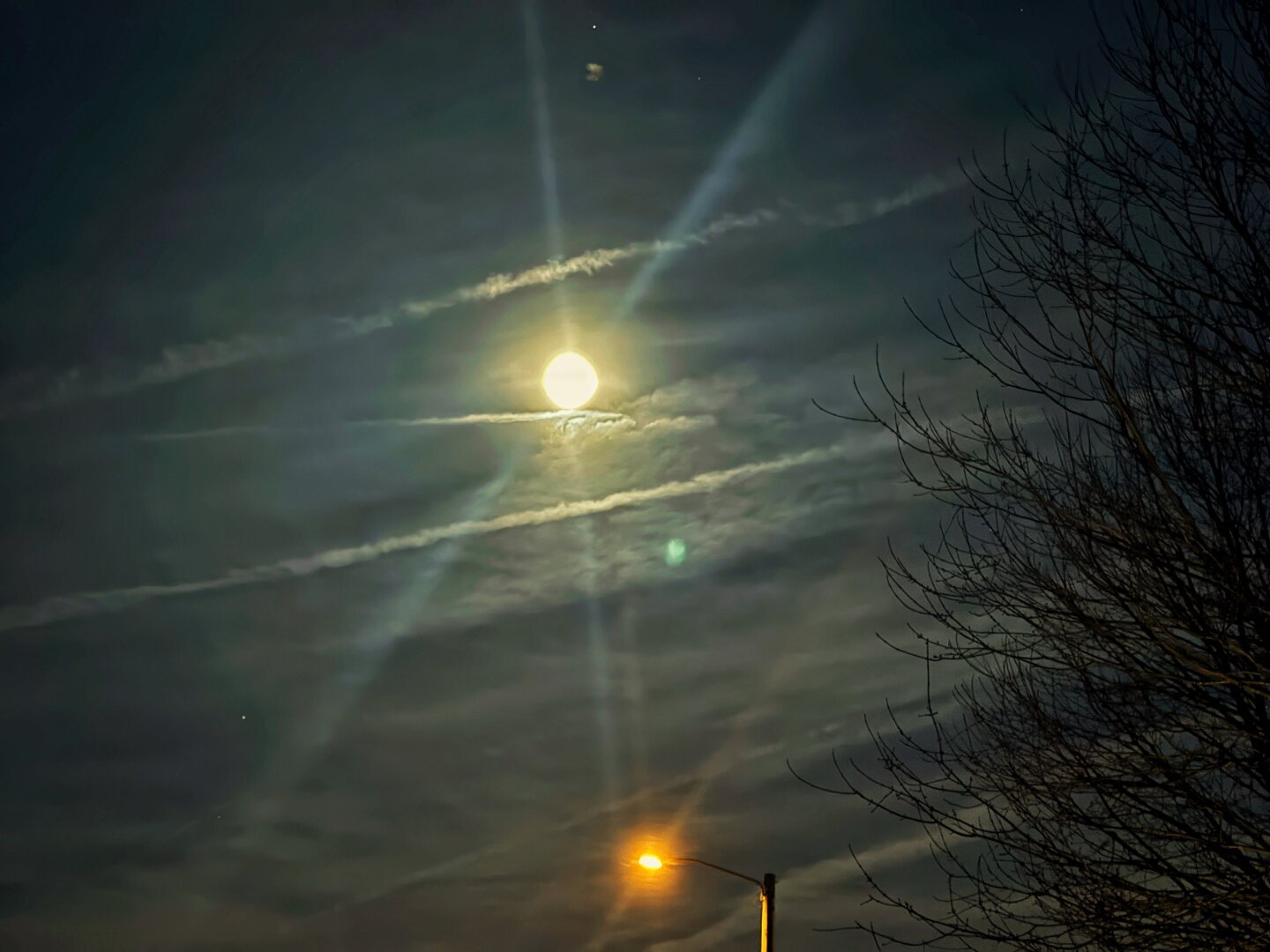 A nighttime shot of the sky contains a bright moon that is surrounded by thin clouds that are horizontally stretched. There are contrails that are visible and go from left to right. In the bottom of the image, there is a street light with a yellow glow. In the right of the image, there is a bare tree.