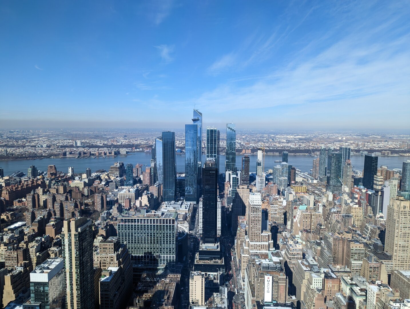 An aerial view of a cityscape with a bright blue sky. The skyline is filled with tall buildings, the tallest buildings are centered in the image. The buildings are a mix of modern and older styles. There is a river in the background. The sky is clear and blue with a few clouds.